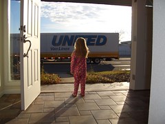 young girl in footie pajamas looks out the front door at a huge moving truck