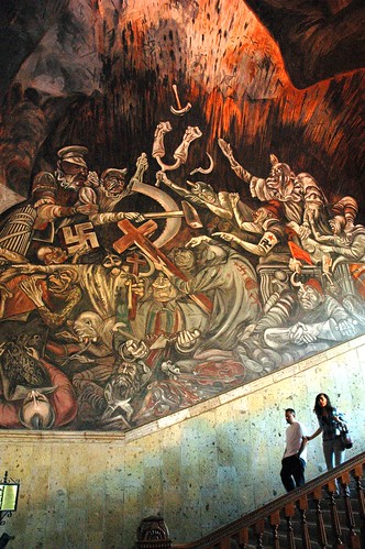 Scale of the large and overwhelming murals, a couple descending the staircase, The Clowns of War Arguing in Hell, depicting war mongers with their political agendas and logos, José Clemente Orozco Mural, Governor's Palace, Guadalajara, Jalisco, Mexico by Wonderlane