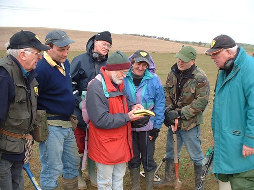 A group of people standing together in a field. They are being shown by a Finds Liaison Officer how to use a GPS device to get a grid reference.
