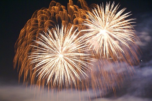 Image shows a huge background burst of copper-colored sparks, with two blooms of bright off-white in front. Smoke drifts like thin cirrus clouds in front towards the bottom and the right of the photo.