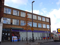 A rather blocky three-storey brick building with dustsheets over the ground floor windows.  An entrance to the upper floors is visible on the left, with the words “Premier House” above.  A Lycamobile-branded phone accessories stall is next to this.