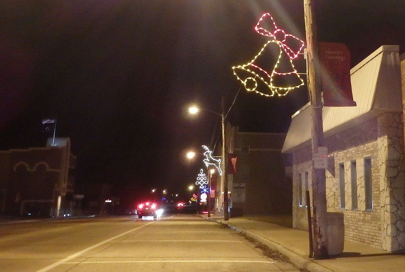 lighted set of bells, reindeer, and curly tree one one side of a busy street