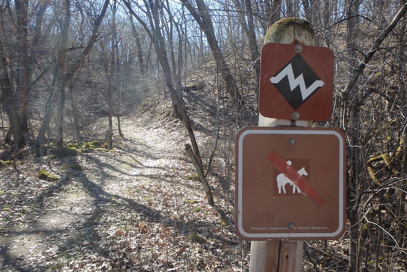 a leaf-covered path at the left, a sign showing a steep incline and another sign forbidding horses