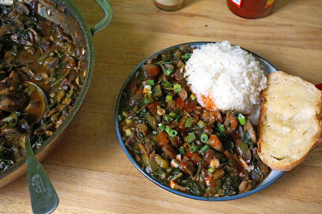 A plate of smothered mushrooms and spinach, all dressed up with hot sauce and green onions, accompanied by rice and bread. A big deep skillet of the dish sits next to the plate