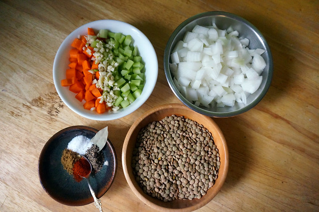 On a wooden counter, bowls of onion, carrots and celery, lentils, and spices. Other than oil and water, this is literally everything that makes lentil soup