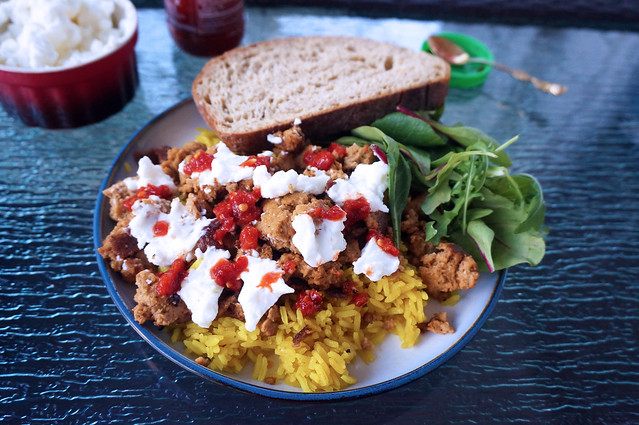 A plate full of street-cart seitan and yellow rice, dotted with garlic sauce and chile paste, a small pile of salad and a slice of bread at the edge of the plate. In the background, a ramekin of garlic sauce, the bottle of chile paste, and the lid to that bottle, with a small gold-plated spoon resting in it.