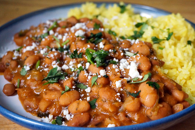 Closeup on pinto beans stewed with cilantro and bay, with light yellow rice in the background; the whole plate is strewn with chopped cilantro and crumbled cotija cheese