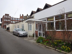 A slantwise view of a long building with a covered porch, seemingly of multiple architectural styles.  Three gable ends are visible above the ground floor.  A small garden grows in front, and a car is parked outside.