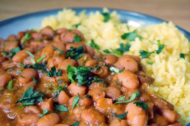 A closeup of pinto beans stewed with cilantro and bay, flecked with bits of chopped cilantro sprinkled over the top. Light yellow rice sits alongside the beans, in slightly unfocused in the background.
