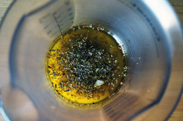 A camera angle looking down into an a blending jar: golden olive oil, on top of which rest gray-green oregano flecks and chunks of cream-colored garlic