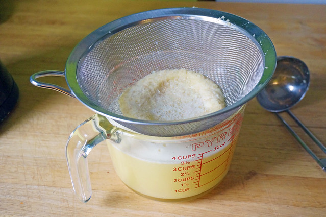 A fine-mesh strainer rests over a measuring cup full of pale, cloudy yellow liquid. In the strainer, a ring of ginger pulp.