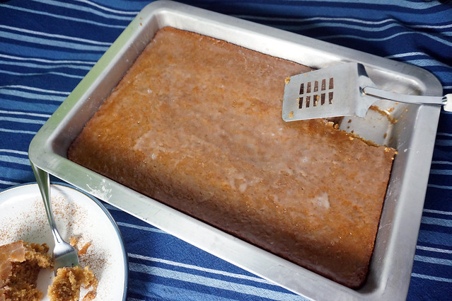 A pan of No-Cola cake sits on a blue striped tablecloth. One piece is missing from the corner of the pan, and a metal spatula rests in the hole where it used to be. Part of the missing square is visible, on a plate, in the lower corner.