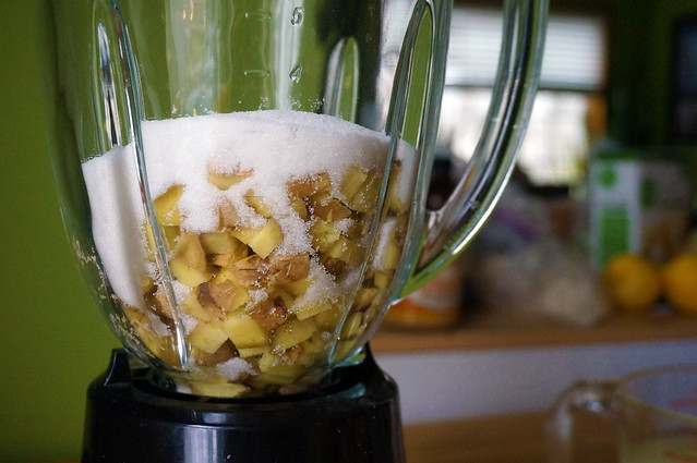 Closeup on chopped ginger in a blender jar, tiny crystals of sugar spilling down among the chunks. A crowded countertop, full of bags, boxes, lemons and limes, is blurred in the background.
