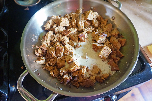 Chopped, pan-fried seitan fills a large, two-handled skillet on a black stovetop; a pool of pale-yellow marinade in the middle waits to be stirred in.