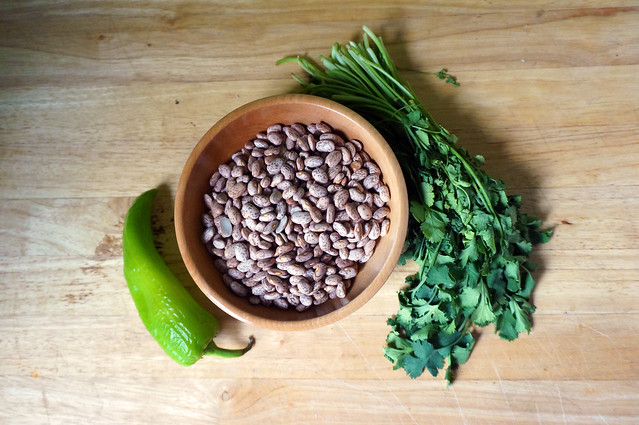 A small wooden bowl filled with pinto beans, flanked by a light green Anaheim pepper and a dark-green bunch of cilantro: yeeeeesss