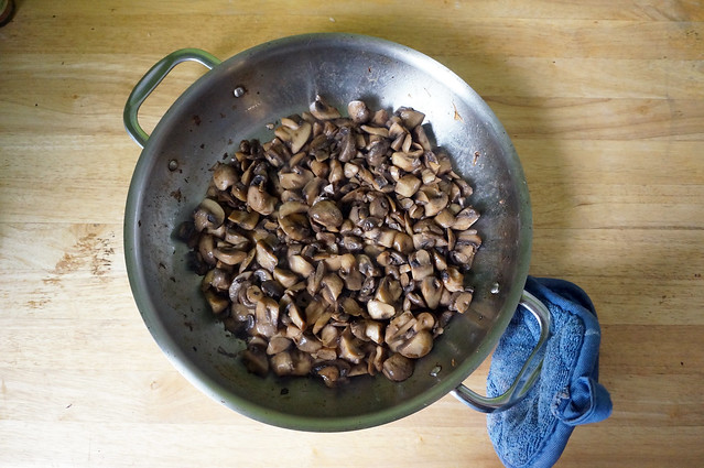A skillet full of chopped mushrooms, wilted and browned, sits on a wooden countertop. A blue hotpad rests on one of the skillet's loop handles