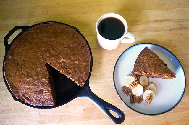 An overhead shot, with a skillet full of rum & coffee banana nut bread to the left, a plate with a wedge of the bread to the right, and an ivory mug filled with black coffee in between