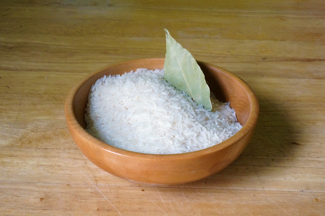 A small wooden bowl filled with raw white rice, with a pale gray-green bay leaf sticking out from the rice. It feels like a Zen garden.