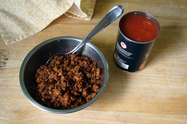 A small metal bowl of cooked chorizo sits on a wooden countertop next to an open can of diced tomatoes