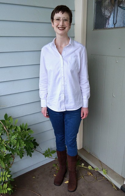 A woman stands in front of blue weatherboard. She wears a white button up shirt and blue jeans.