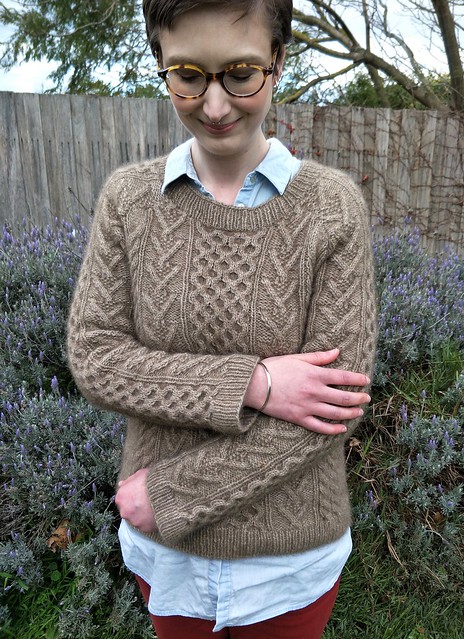 A woman stands in front of a garden fence. She holds her arms to show off the detailed work of her handknit cabled jumper.