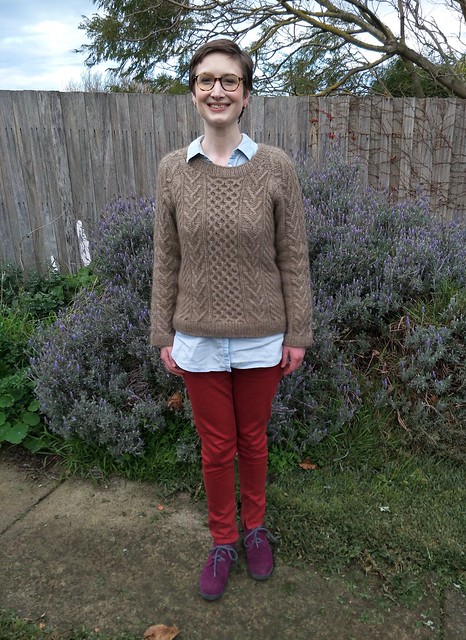 A woman stands in front of a garden fence. She wears a handknit cabled jumper, denim shirt, red jeans and purple suede boots. She is smiling.