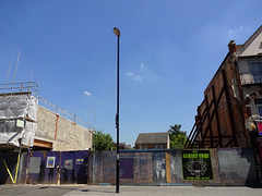A gap in a row of terraced buildings.  On the right is a three-storey “end” of terrace, shored up with props; in the middle is a gap behind worn low hoardings; and on the left is the beginnings of a rebuilt part of the terrace, still with scaffolding on.