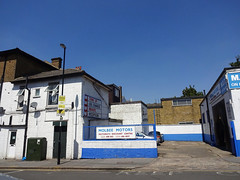 A photo taken on a sunny day with a cloudless blue sky, showing a yard between white-painted buildings to left and right, with exposed-brick buildings in the background.  A couple of cars are parked in the yard, and a sign on the wall at the front reads “Molbee Motors / Motorists Discount Centre”.  Two side entrances can be seen to the building on the left; one a modern white PVC door and the other covered with a dark grey pull-down shutter.