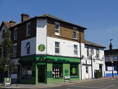 An oblique view onto an end-of-terrace building — three stories at the corner reducing to two stories at the back on the side road.  The ground floor at the corner is painted green with stylised “JC” initials on a window decal and above the corner entrance door.