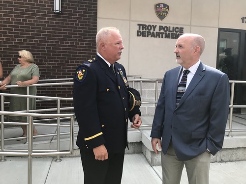 Captain Rick Sprague speaking with Mayor Patrick Madden in front of Troy Police Department headquarters. The Troy Police Department logo is visible on the concrete wall behind Captain Sprague and Mayor Madden