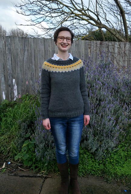 A woman stands in front of a garden fence. She wears a yellow and grey colourwork yoke Icelandic jumper, blue jeans and brown suede boots.
