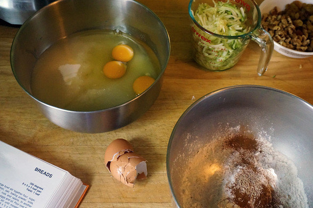 Ingredients for zucchini bread are arrayed in a variety of bowls on a wooden counter; brown eggshells nestle within each other next to an open cookbook