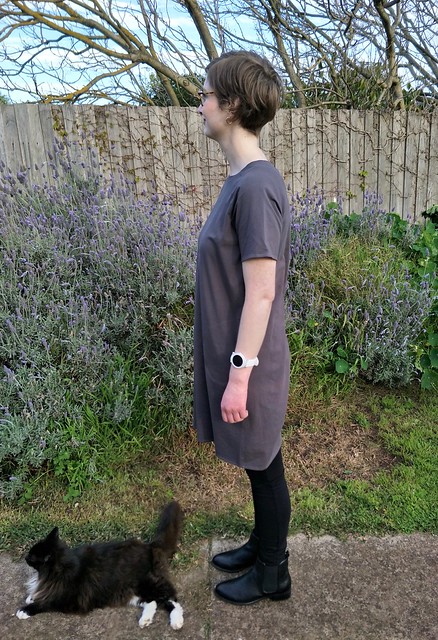 Woman stands in front of garden fence. She wears a short-sleeve, grey knit boxy tee dress with pockets, black leggings and black ankle boots. She is facing side on to the camera, and a black and white fluffy cat rests at her feet.