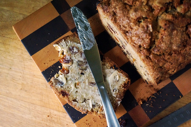 A knife smeared with butter lays across a slice of buttered zucchini bread, which is missing a bite