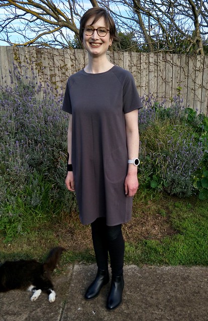 Woman stands in front of garden fence. She wears a short-sleeve, grey knit boxy tee dress with pockets, black leggings and black ankle boots. She is smiling.