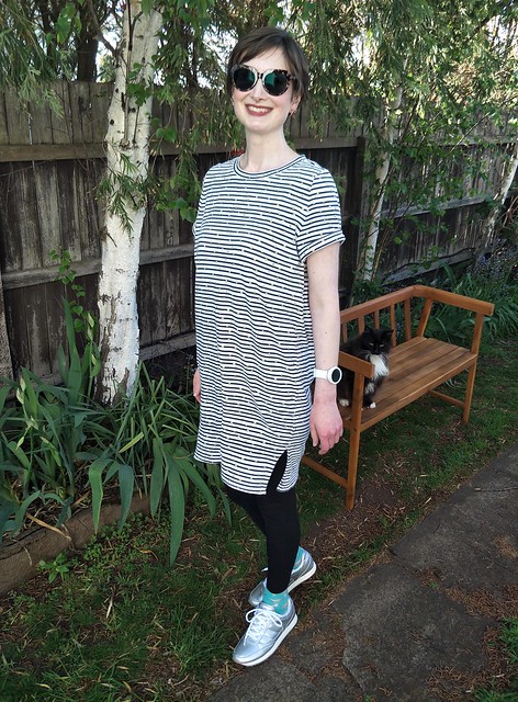 A woman stands in front of a garden fence. She wears a stripe knit tee dress with black leggings, silver runners and big tortoiseshell sunglasses. A cat sits on a bench behind her.