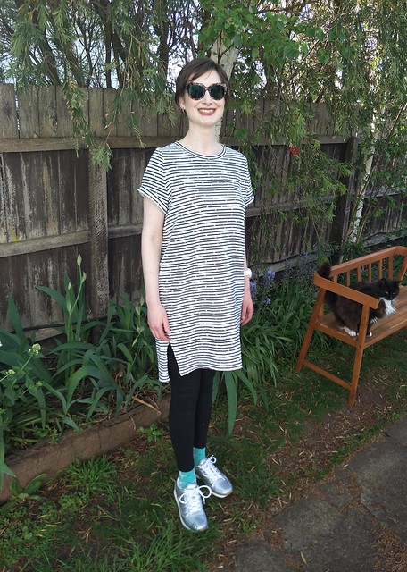 A woman stands in front of a garden fence. She wears a stripe knit tee dress with black leggings, silver runners and big tortoiseshell sunglasses. A cat sits on a bench behind her.